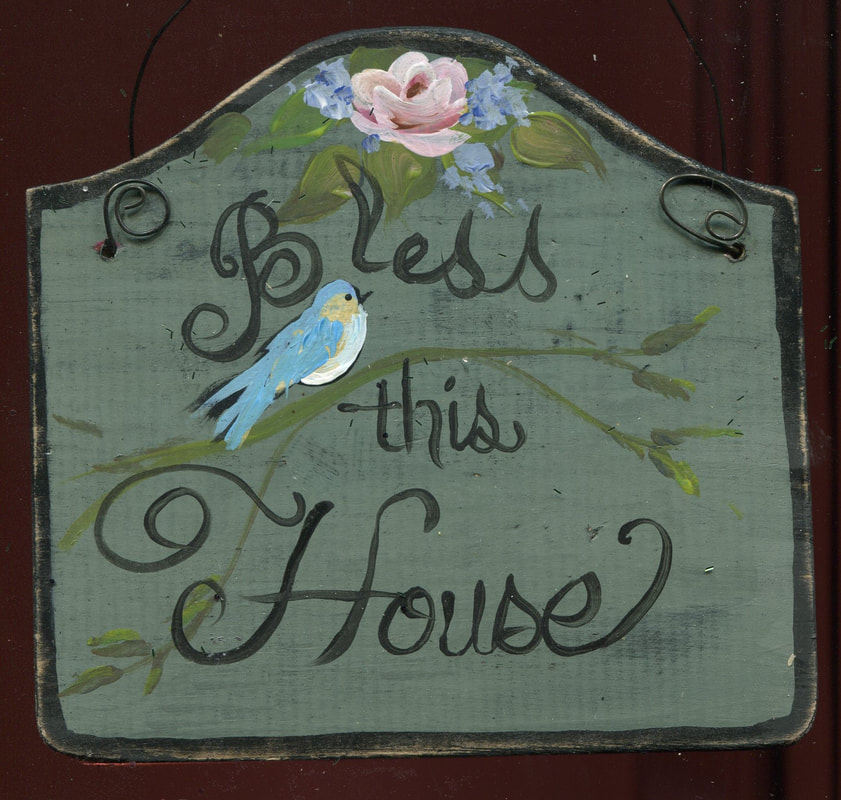 Hand-painted green wood sign reading 'Bless this House' in script, with a bluebird on a branch and a pink rose with blue flowers at the top.